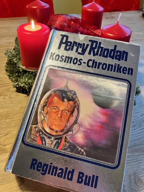 Die Kosmos-Chroniken Band 1 vor einem Adventskranz mit einer brennenden Kerze.